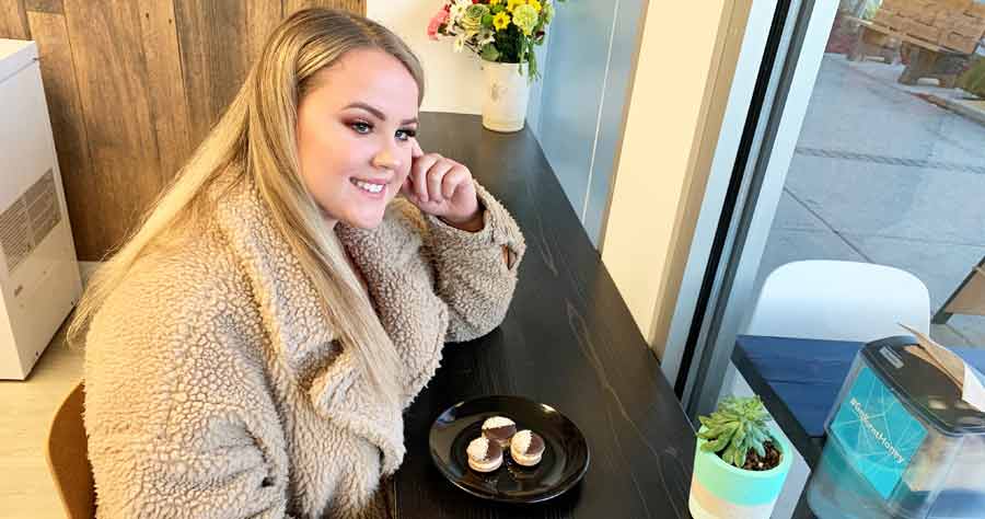 A woman sits at a table, smiling. A plate with a pastry on it is in front of her.