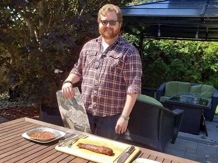 A man stands in front of a table, smiling. Barbecued meat sits on a plate.