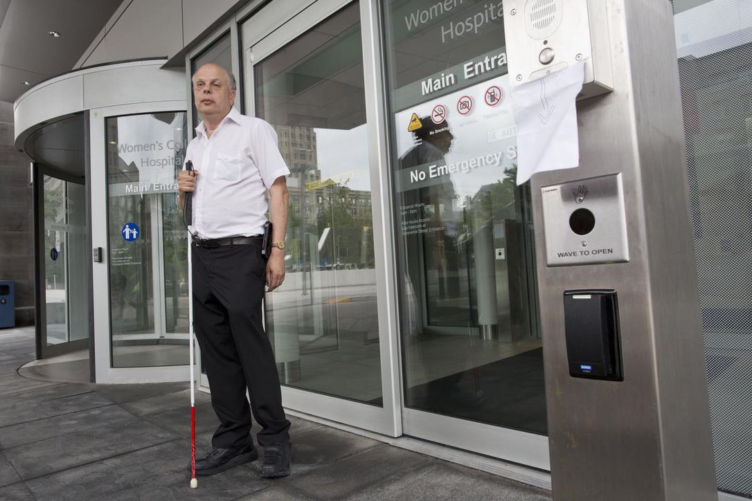 A photo of David Lepofsky with a white cane in front of hospital doors. 