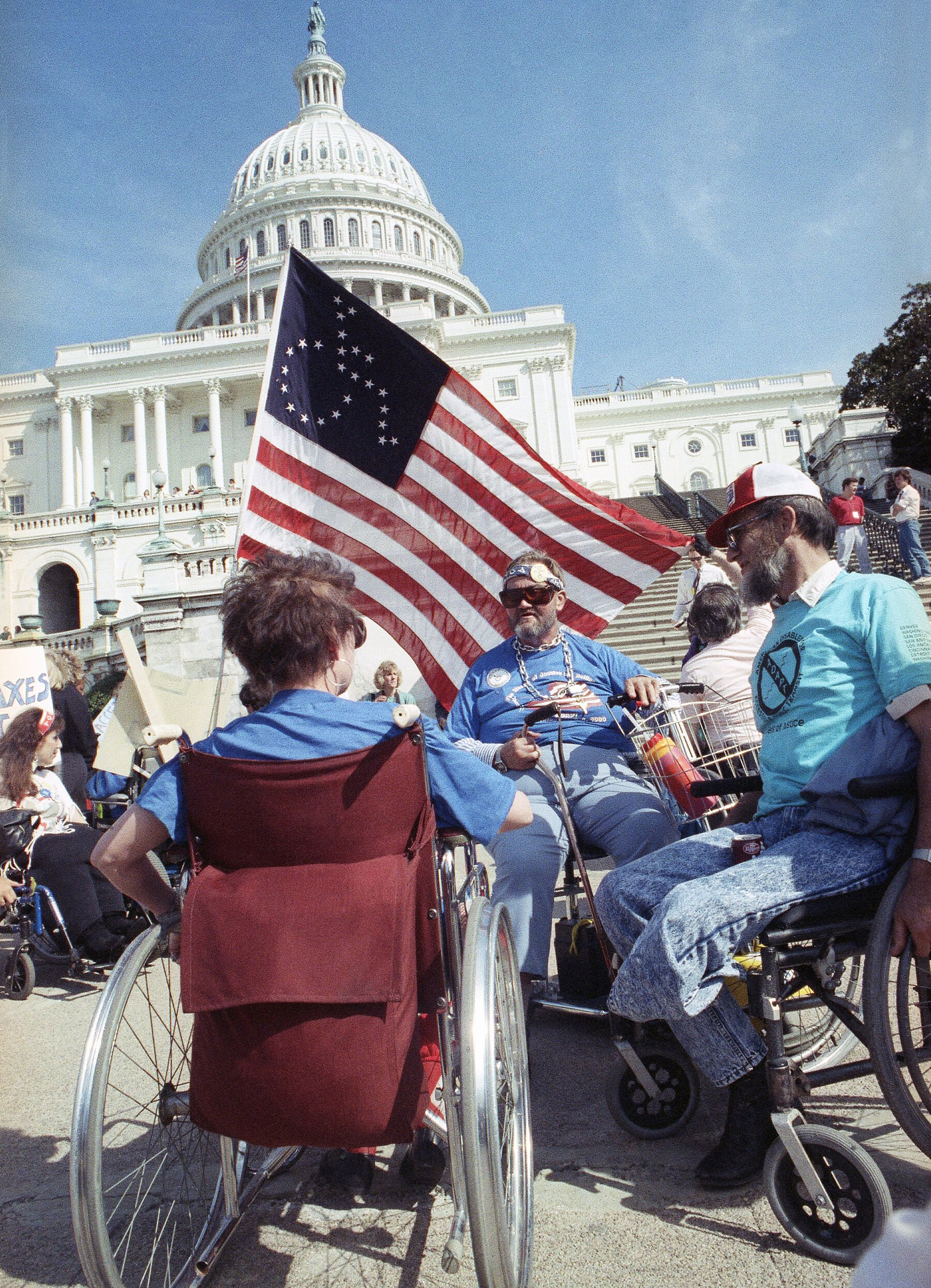 A group of people in wheelchairs outside the US Capitol building holding an American flag with stars that form the universal symbol of access.