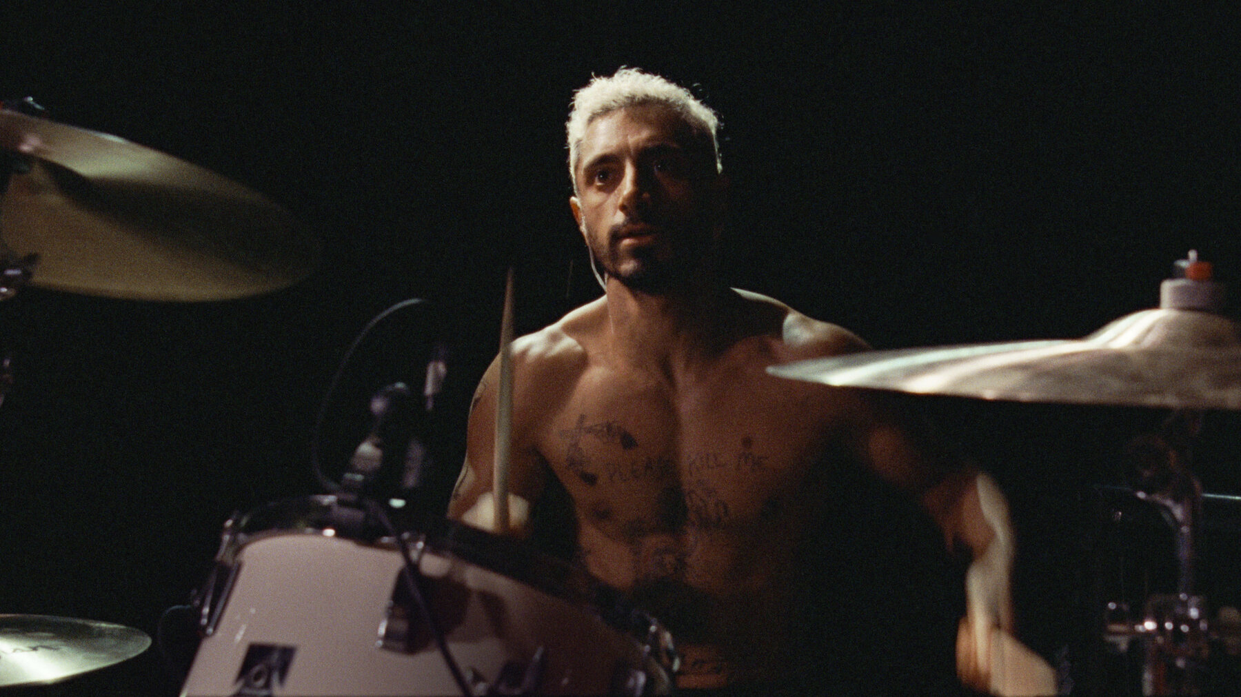 A shirtless Riz Ahmen in character sitting at a drumset.