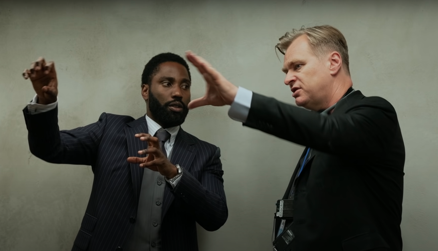 A photo of John David Washington on set and in character with director Christopher Nolan. Both are chatting with their hands in the air. 