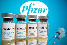Four vials of COVID-19 vaccine and a syringe.  The word Pfizer is displayed in the background.