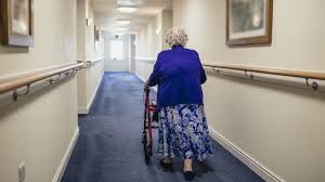 Elderly woman walks in hallway using walker.