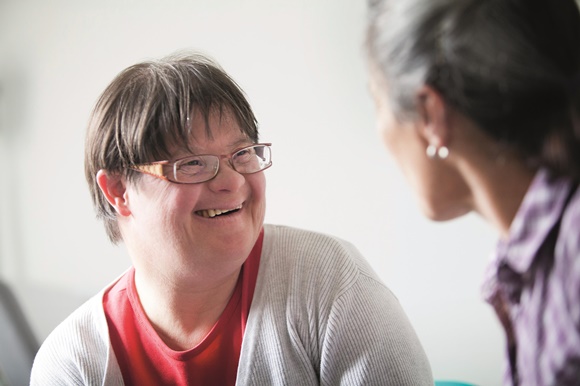 An adult woman with down syndrome smiles at another woman.