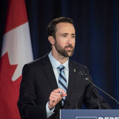 A photo of Derek Sloan wearing a suit and speaking into a podium microphone in front of a Canadian flag.