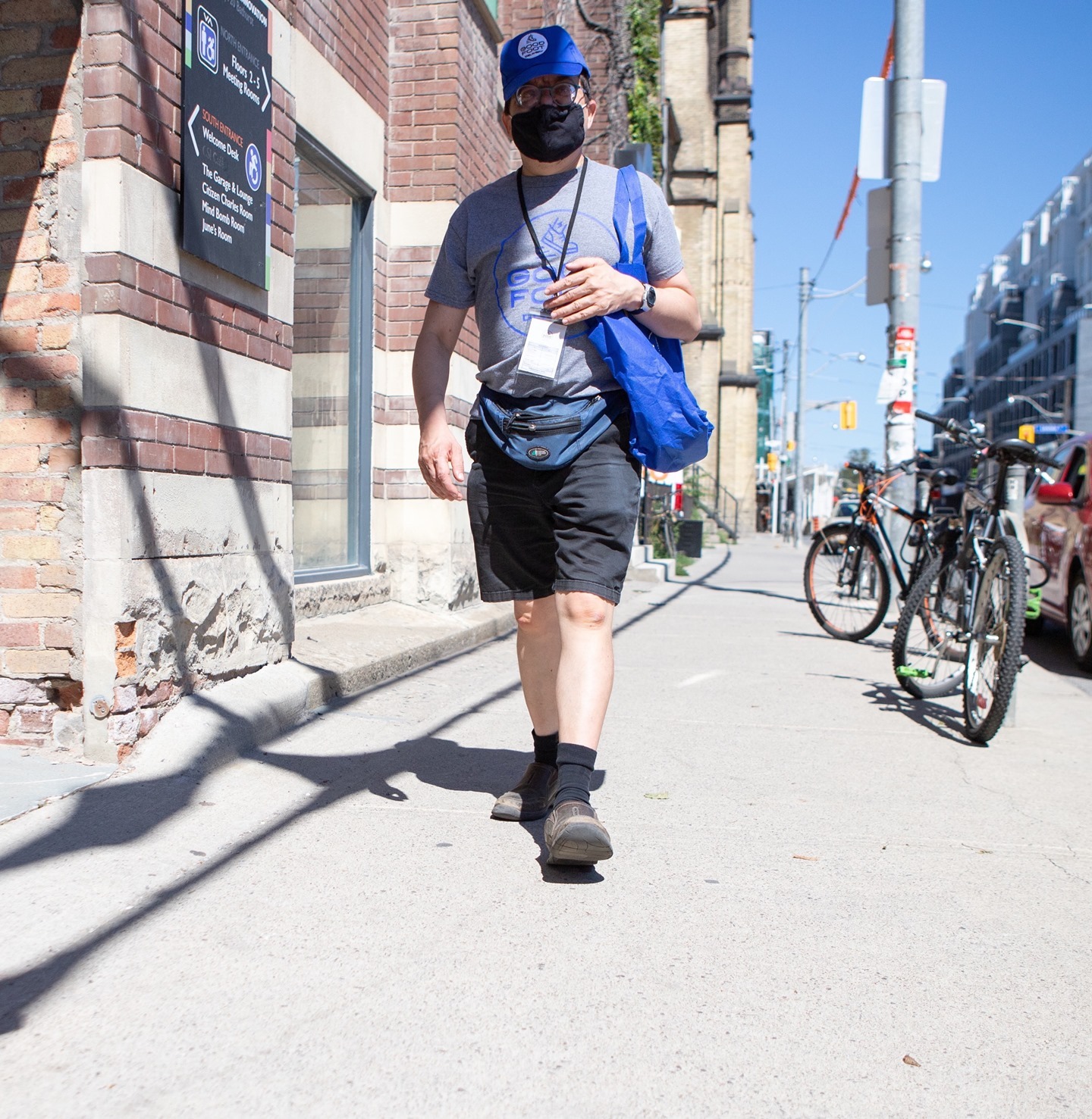 A man wearing a mask walks down the sidewalk holding a blue Good Foot delivery bag. 