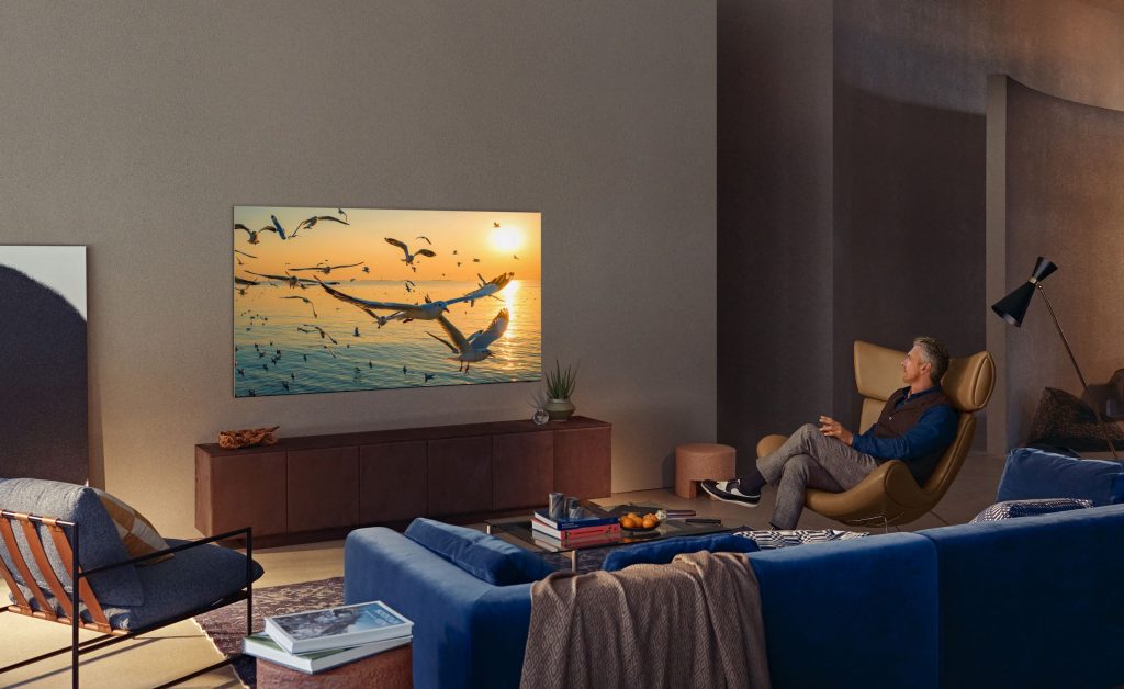 A man sits in his living room watching a Neo QLED, a large Samsung flat screen TV on the wall.