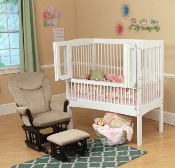 A white baby crib with open bi-fold doors beside a sofa rocking chair. 