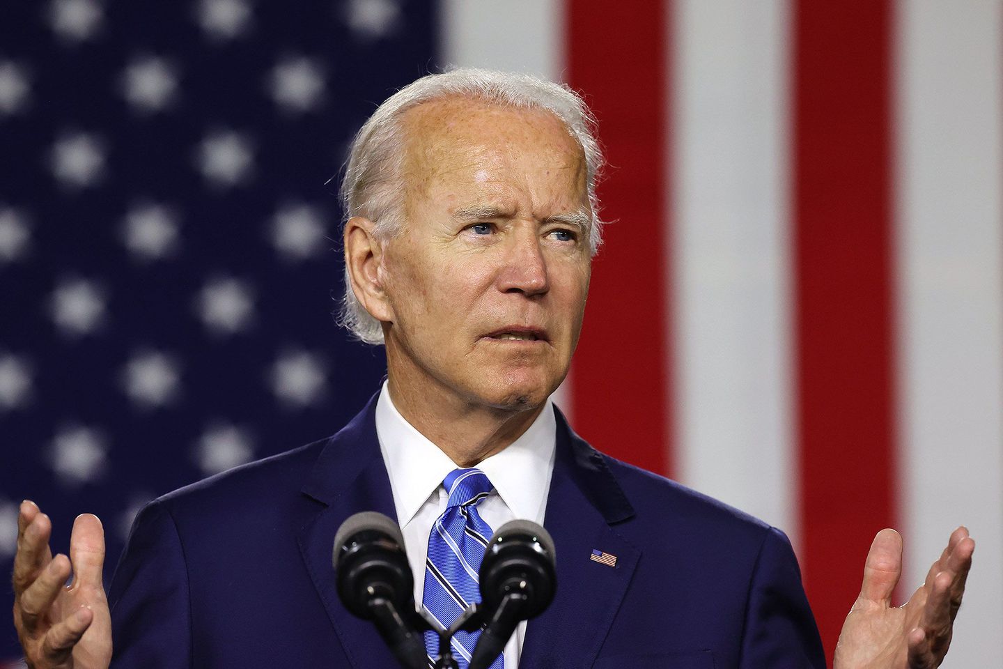 A picture of Joe Biden in a suit speakng at a podium with his hands gestruing. Behind him is an American flag. 