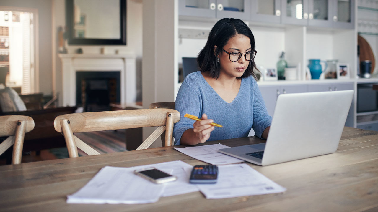 A woman works from home at a desk with a laptop and paperwork. 