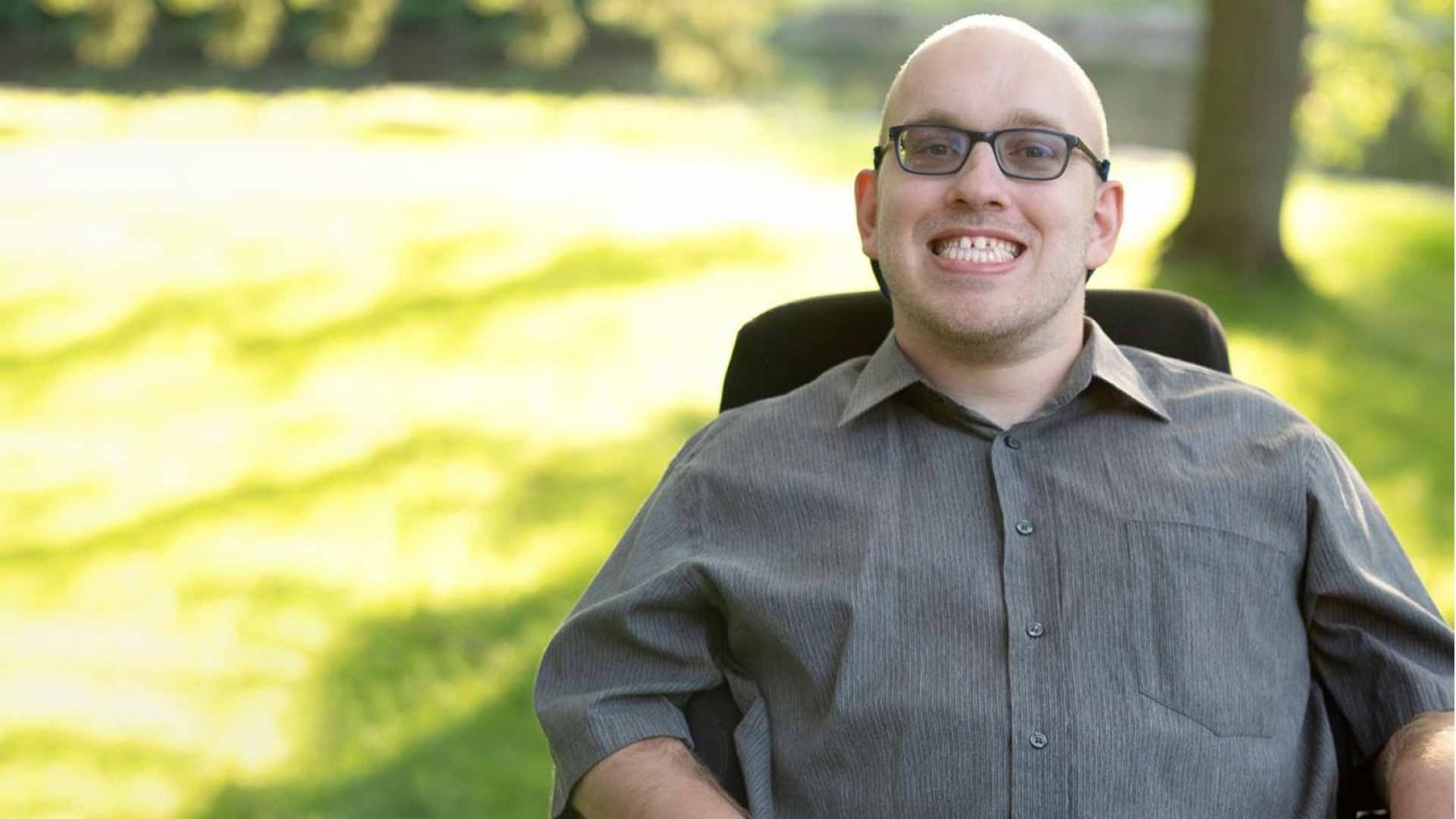 A photo of Brock Richardson smiling in his wheelchair in a green space outdoors. 
