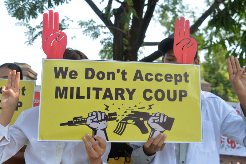 Two protesters are holding up a sign, covering their faces, that reads "We don't accept Military Coup." Their other hands are holding up three fingers. 