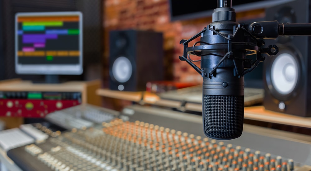 A closeup of a professional radio mic with a mixing board and a computer behind it. 