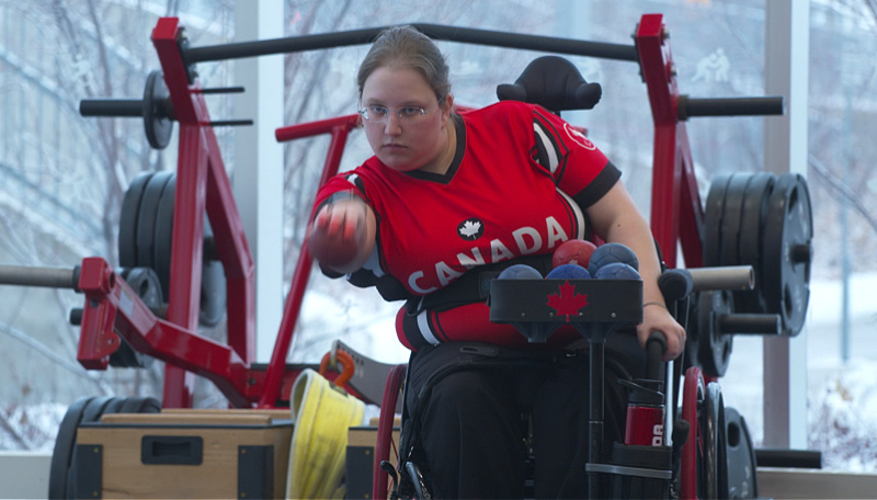 A woman, using a wheelchair, throws a fist-sized ball in a gym.