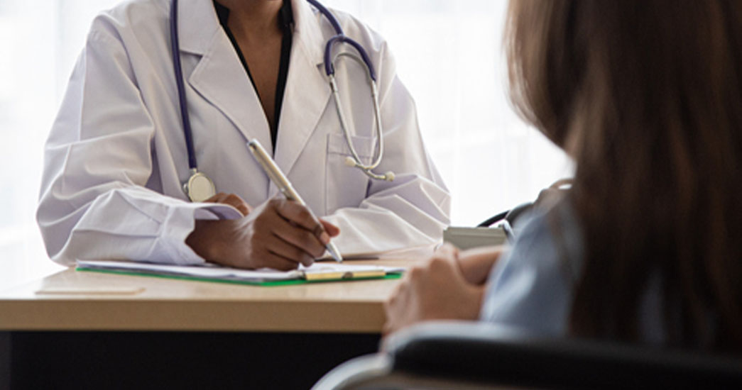 A healthcare worker of colour taking notes at a fdesk in front of a patient who is caucasian. 