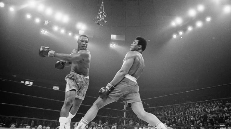 A black and white photo from the 1971 of boxer Joe Frazier swinging a punch at Muhammad Ali who is leaning back and missing the punch. 