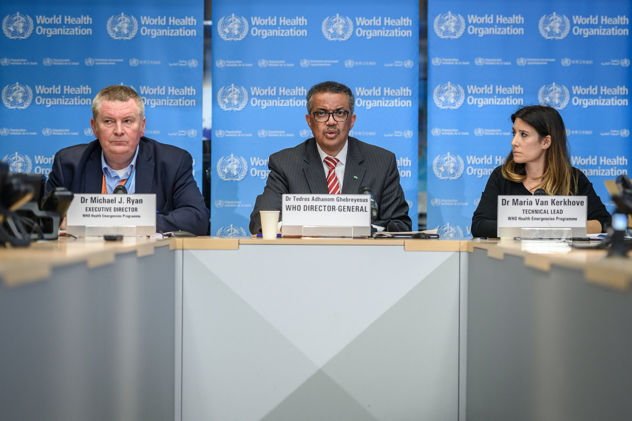WHO chief Tedros Adhanom Ghebreyesus speaking while seated at a press conference table between two other WHO doctors. 
