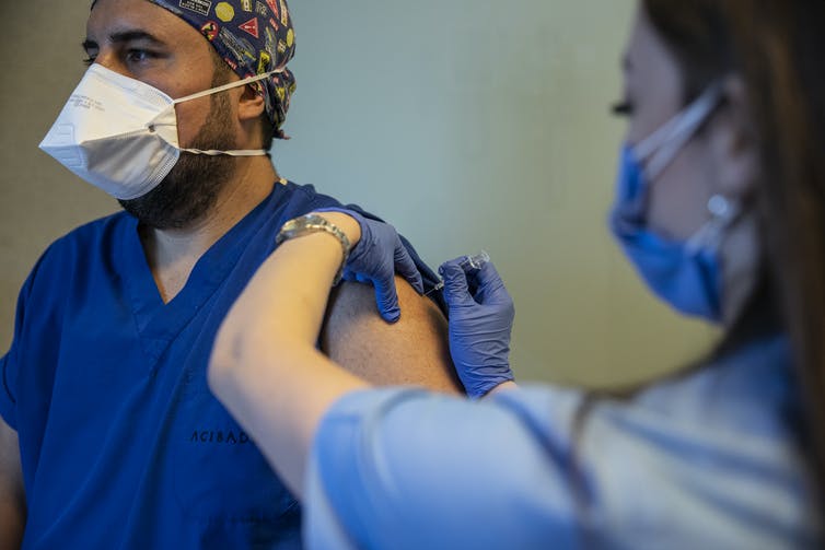 A health care worker gives a vaccine into another health worker's arm. Both are wearing face masks.