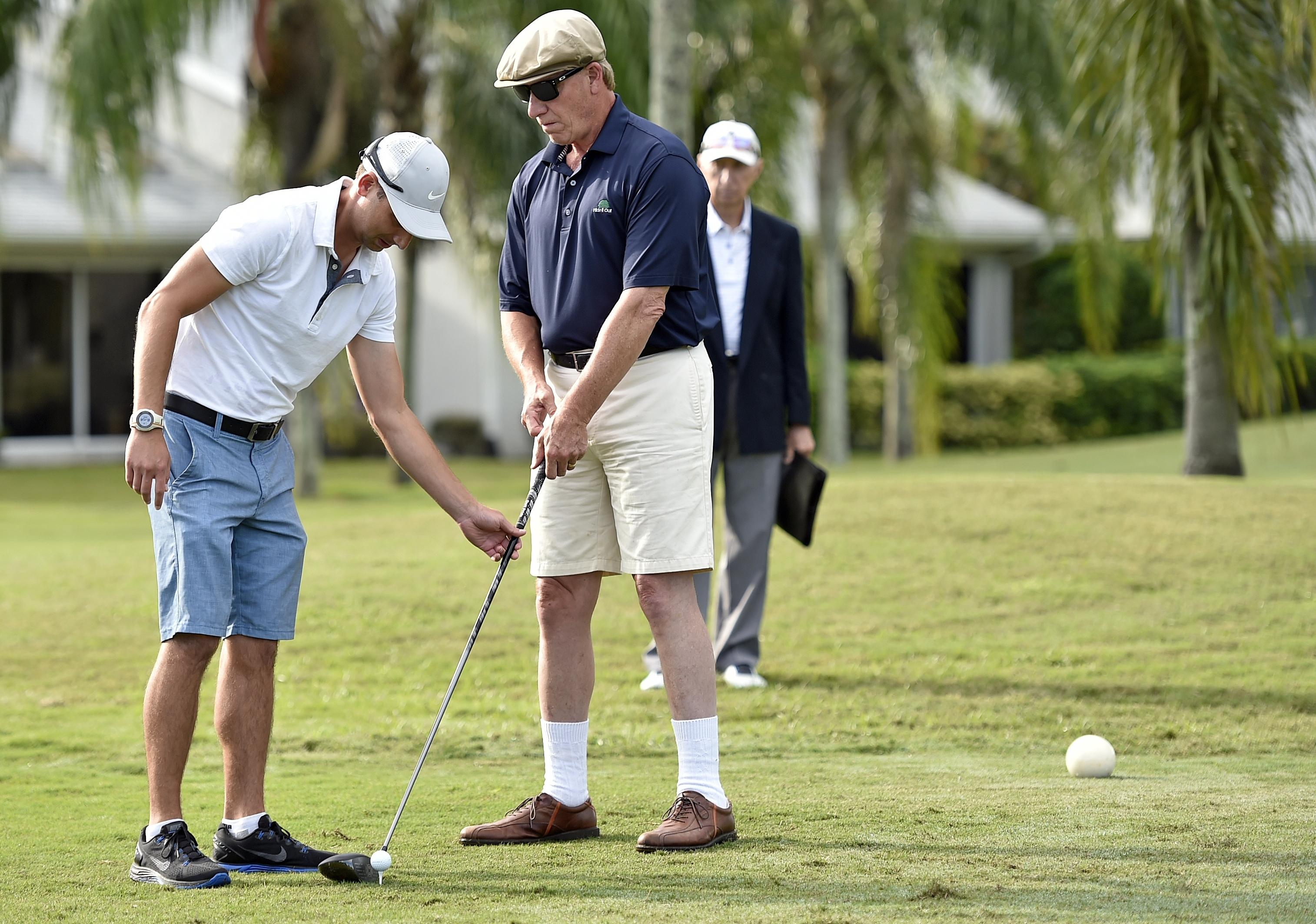 A man assists a blind golfer to align his club to the ball on the ground of a gold course. 