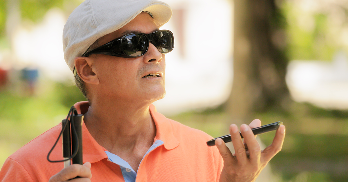 A man wearing sunglasses with his white cane, holds his phone up to listen. 