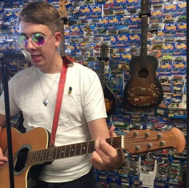 A photo of Justin Wack weaeing sunglasses and playing guitar in front of a mic. 