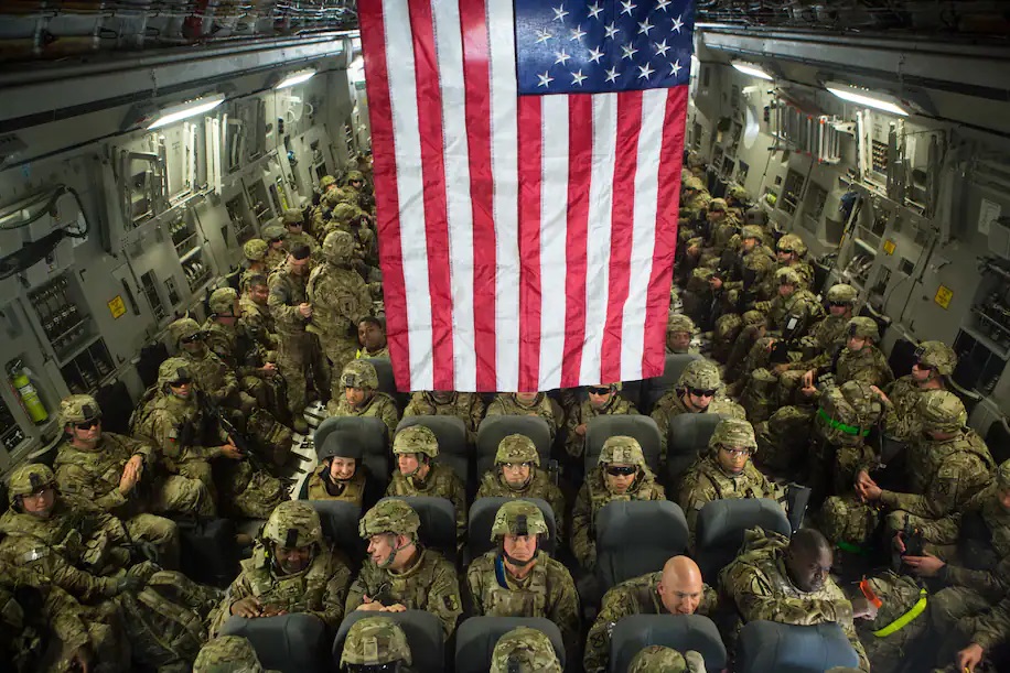 U.S. Troops remain seated in a cargo plane with an American flag hanging above them. 