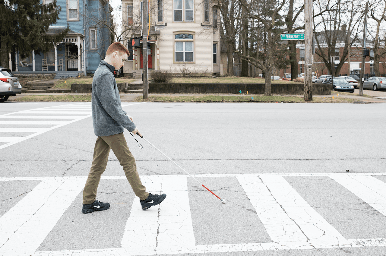 A young man with a white cane crosses the street in a neighbourhood. 