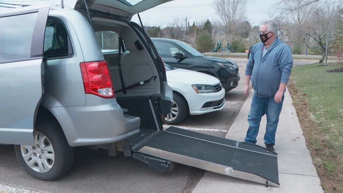 A man with a face mask stands at the back of a van with its trunk open and a ramp sticking out onto the sidewalk. 