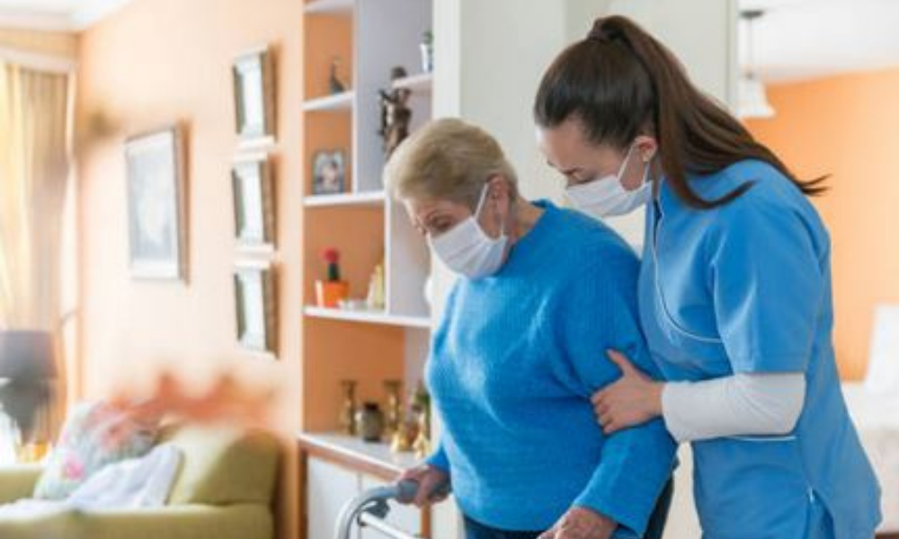 In a living room, a health care worker wearing a face mask holds the arm of an elderly woman wearing a face mask and using a walker. 