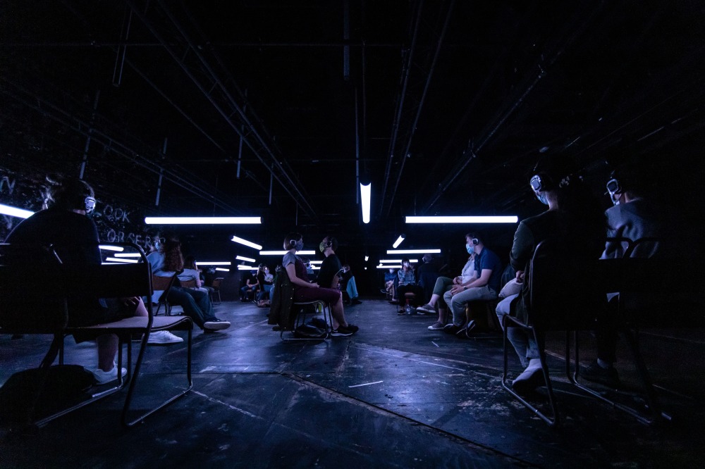 A sound installation photo of the Mirvish Production of Blindness. People sitting on a stage spaced out in the dark wearing headphones and masks.