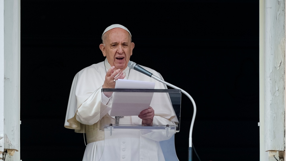 A photo of Pope Francis speaking at a podium mic with a paper in hand.