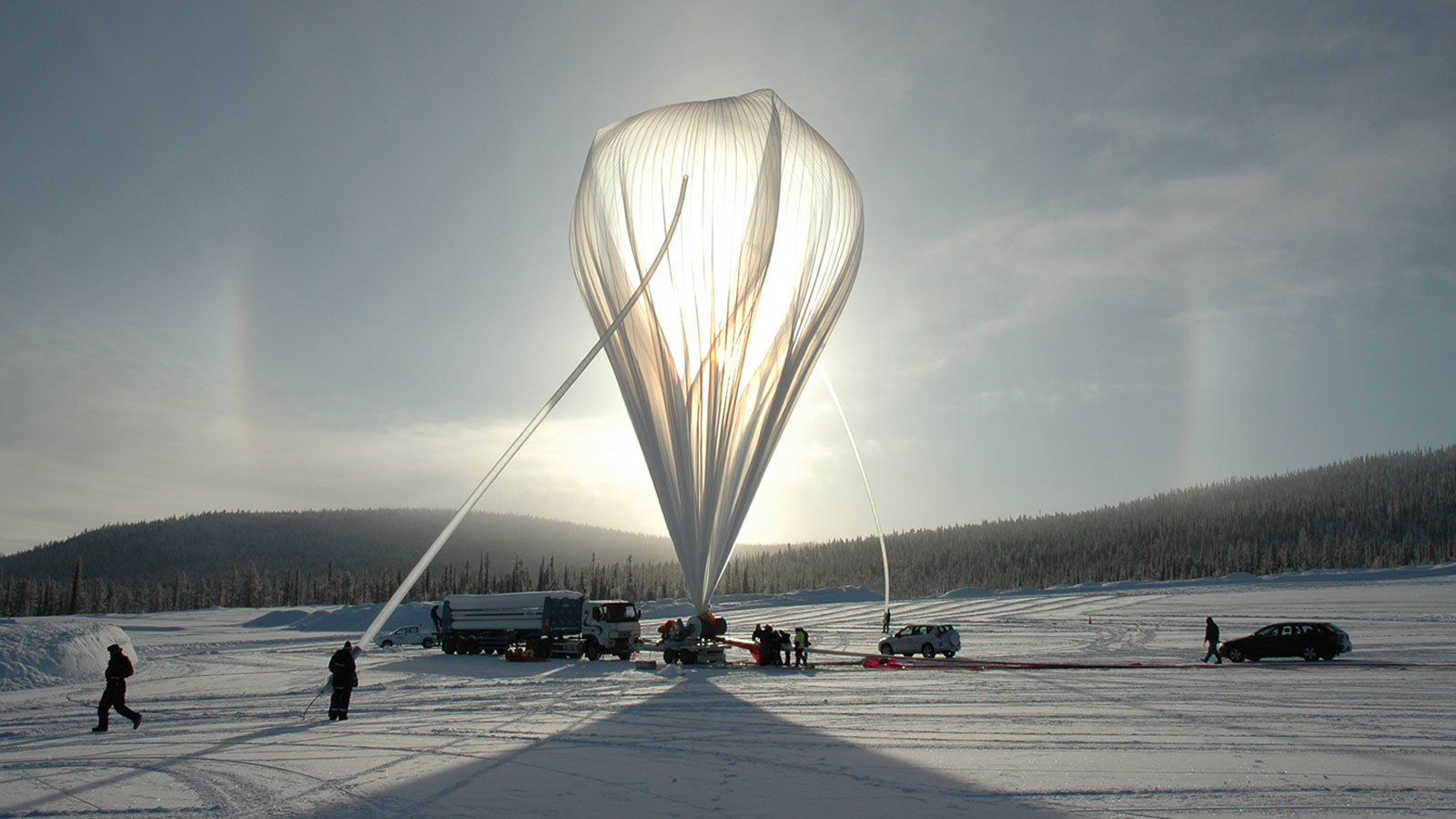 A photo of a massive controlled balloon that would have launched at the Esrange Space Center near Kiruna, Sweden. A few people are on the ground near vehicles. In the distance are forest trees.