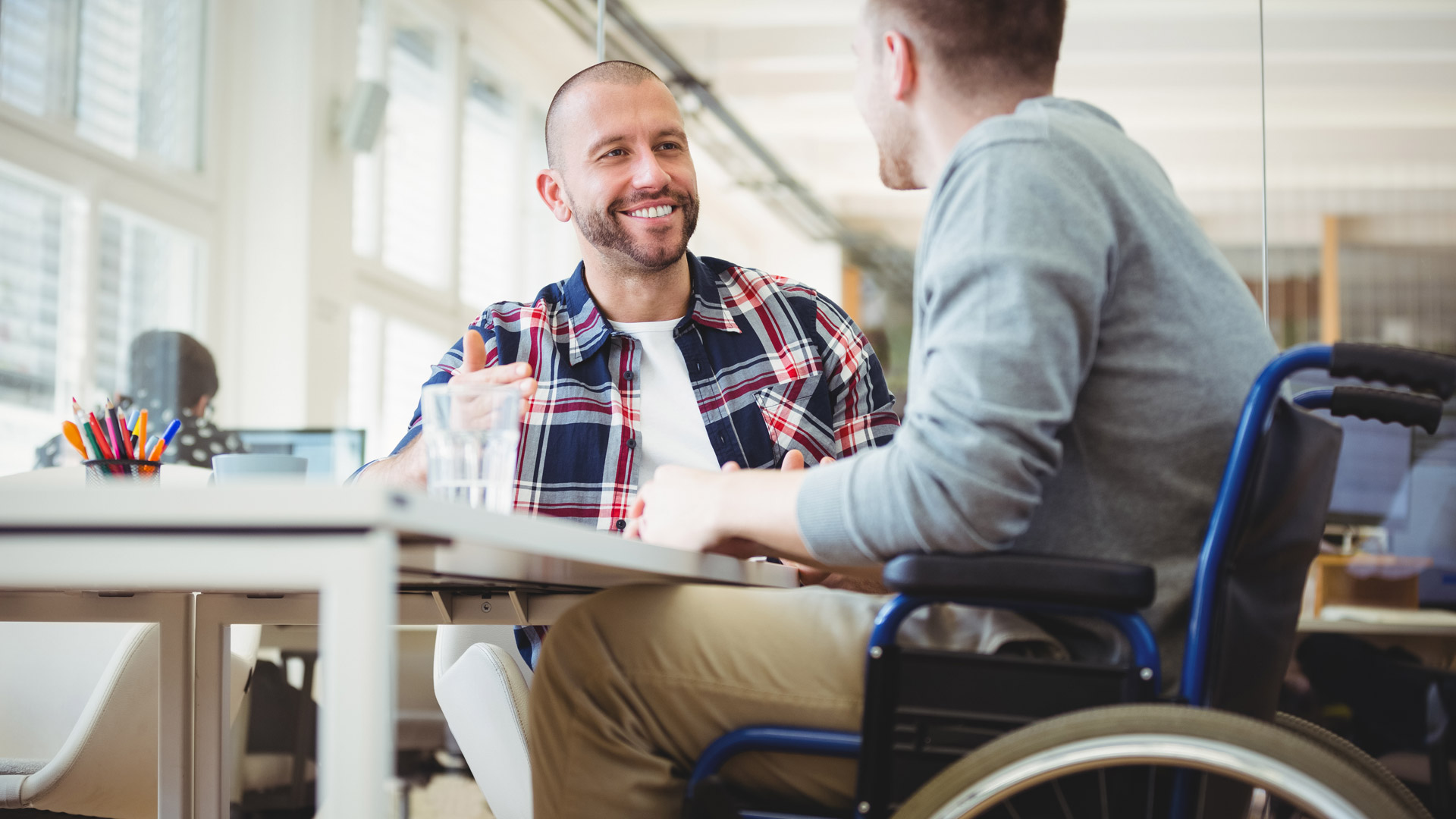 A man in a wheelchair talks with a colleague at a work table. 