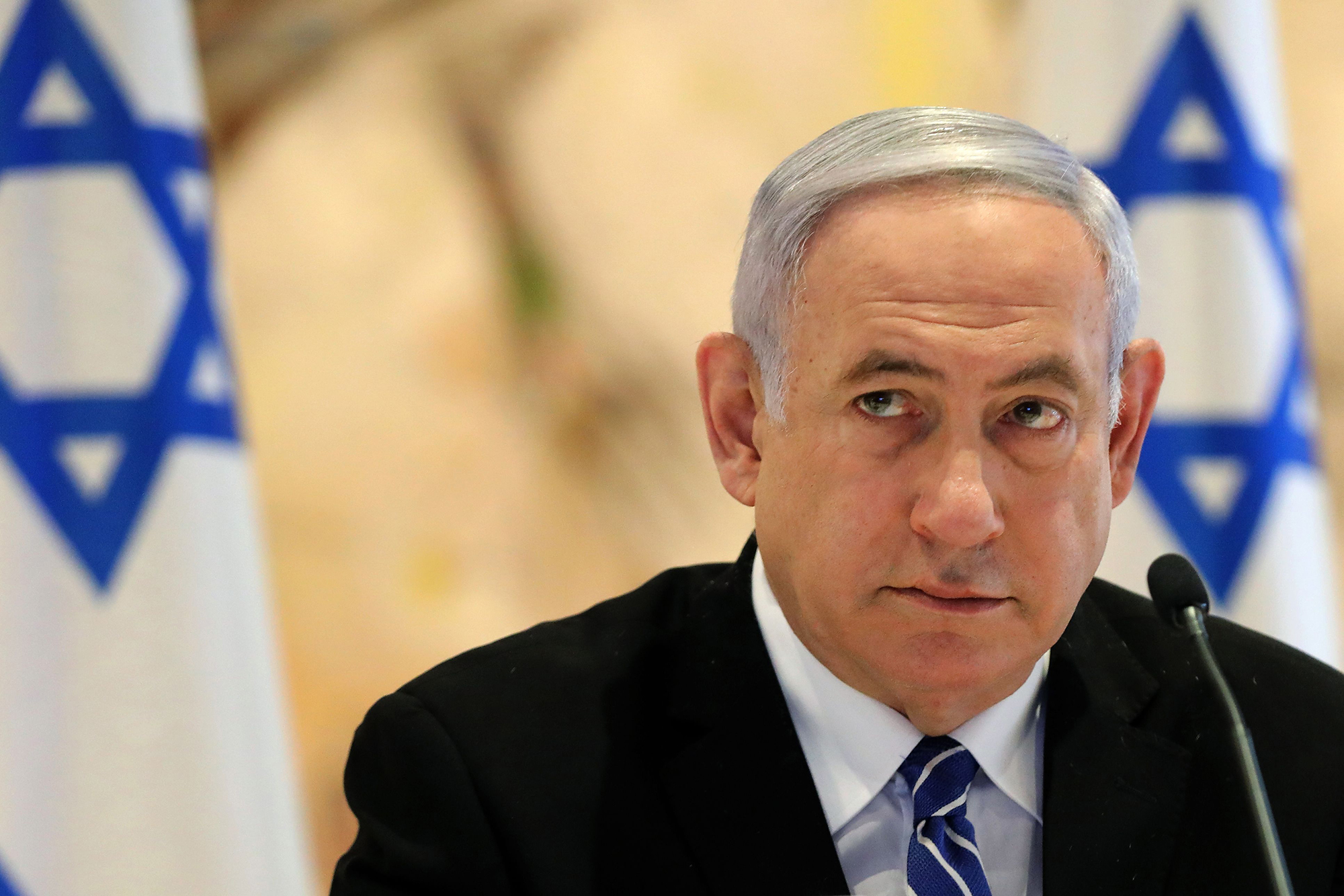 Image shows a close up of Benjamin Netanyahu behind a microphone with two Israeli flags behind him.