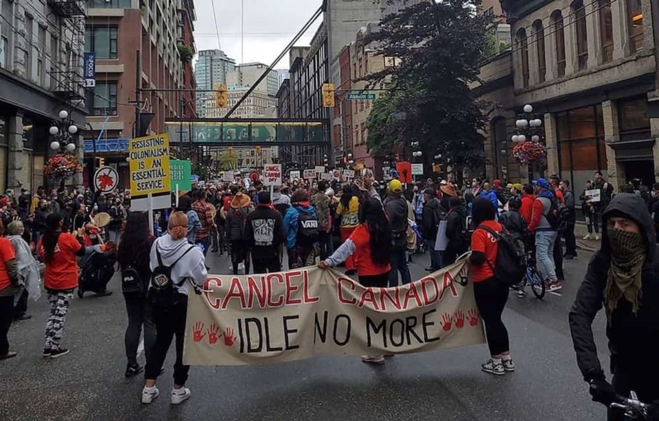 The back of a crowd of people marching with signs. Two people in the forefront hold a long banner that reads "Cancel Canada Day. Idle no more." 