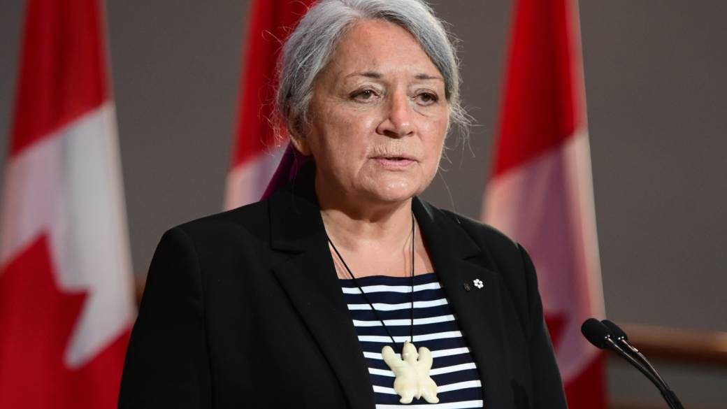 A photo of Mary Simon speaking into a microphone podium with Canadian flags behind her. She is wearing a necklace with a large white turtle pendant. 