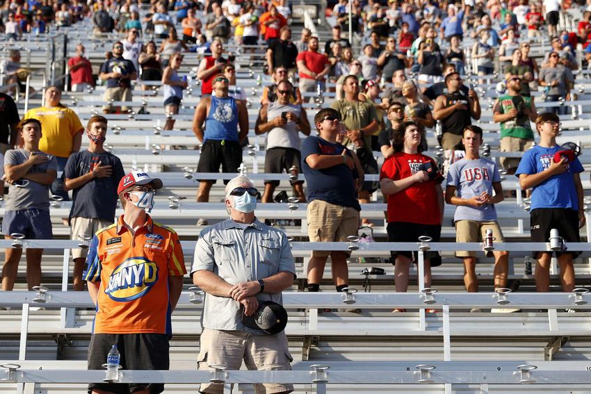 Fans stand spaced out between the outdoor bleacher benches. Some are wearing masks, others are not. 