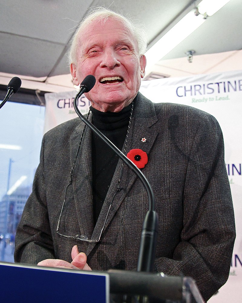A photo of Bill Davis smiling and wearing a poppy while speaking into a microphone. 