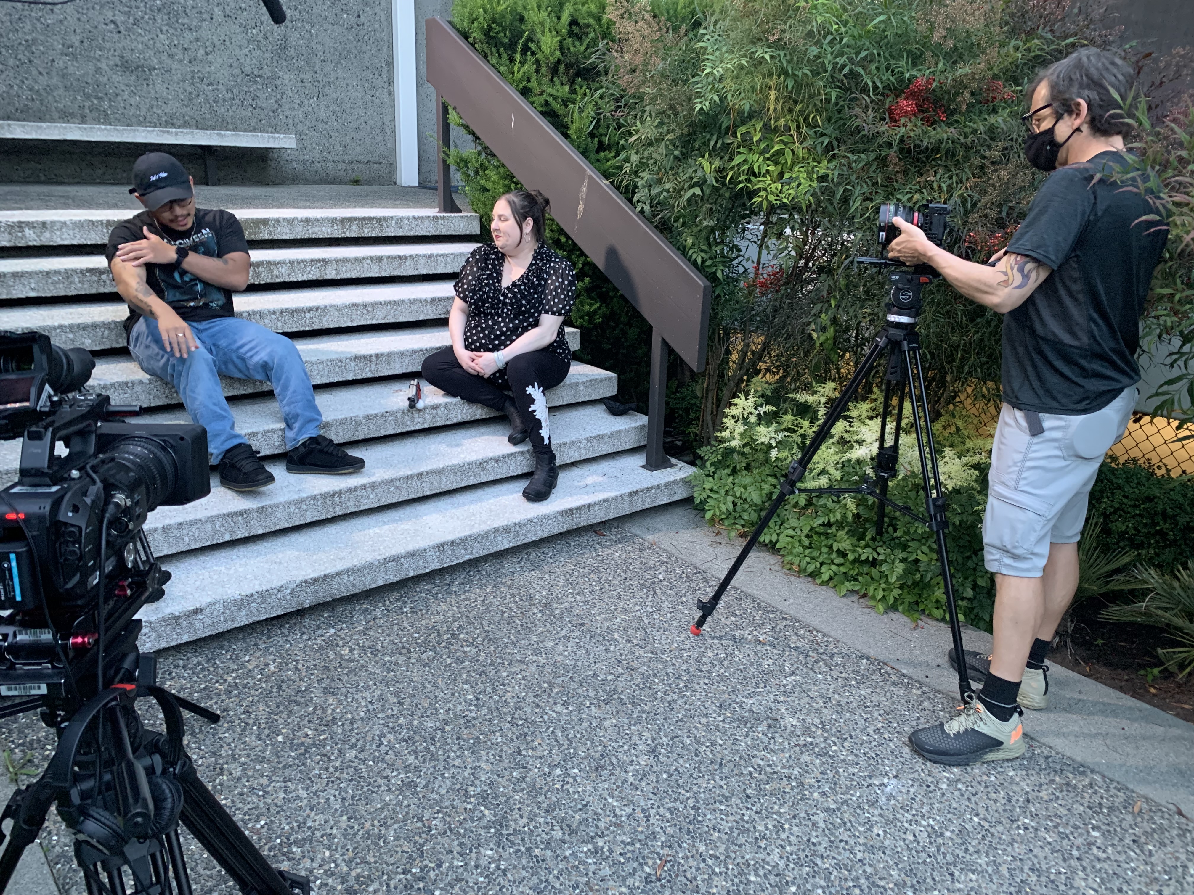 A photo of Amy Amantea sitting outside on a step speaking to a man. A camera crew films them. 