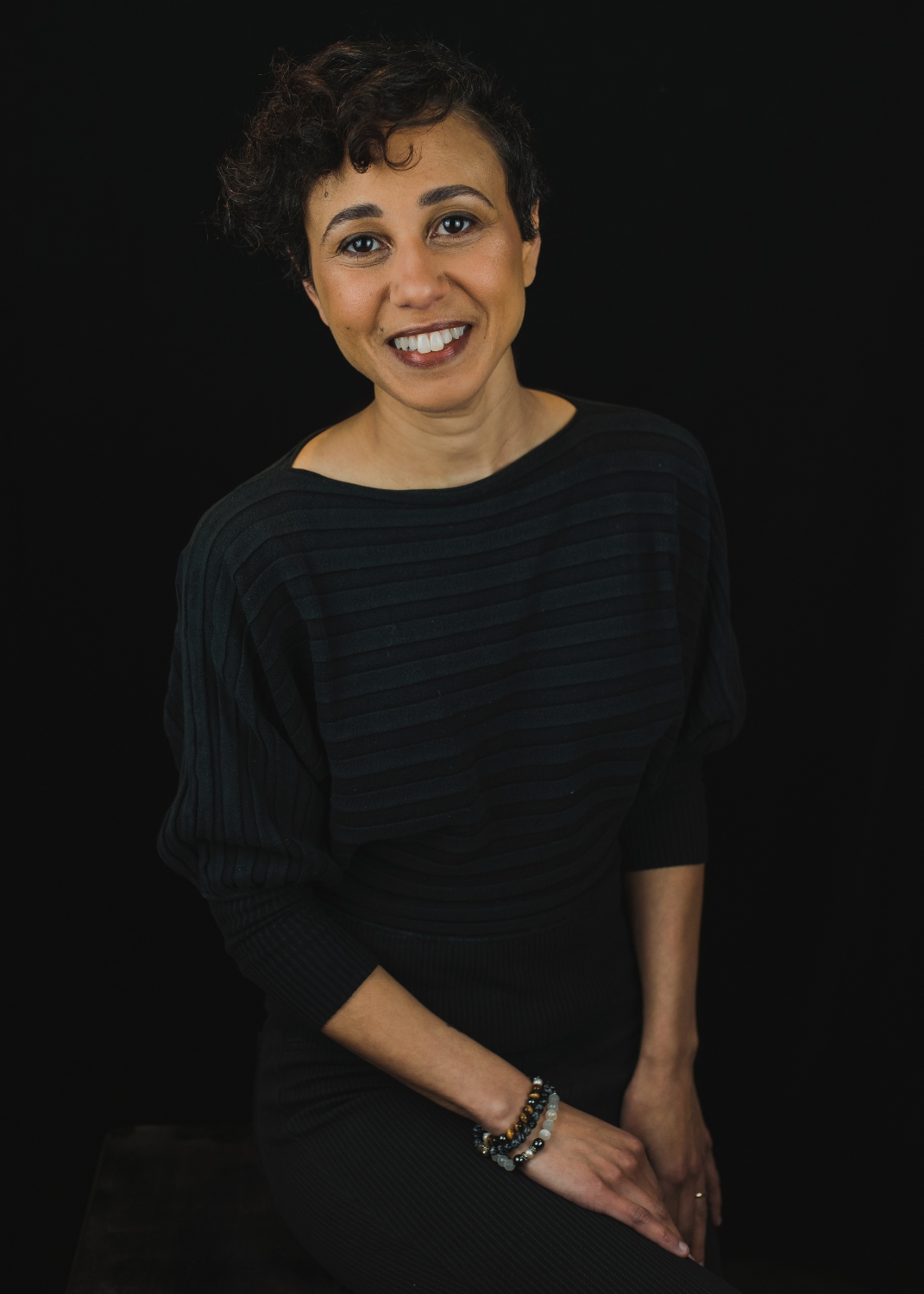 A photo of Kelly Bron Johnson smiling with a dark shirt and a black background. 
