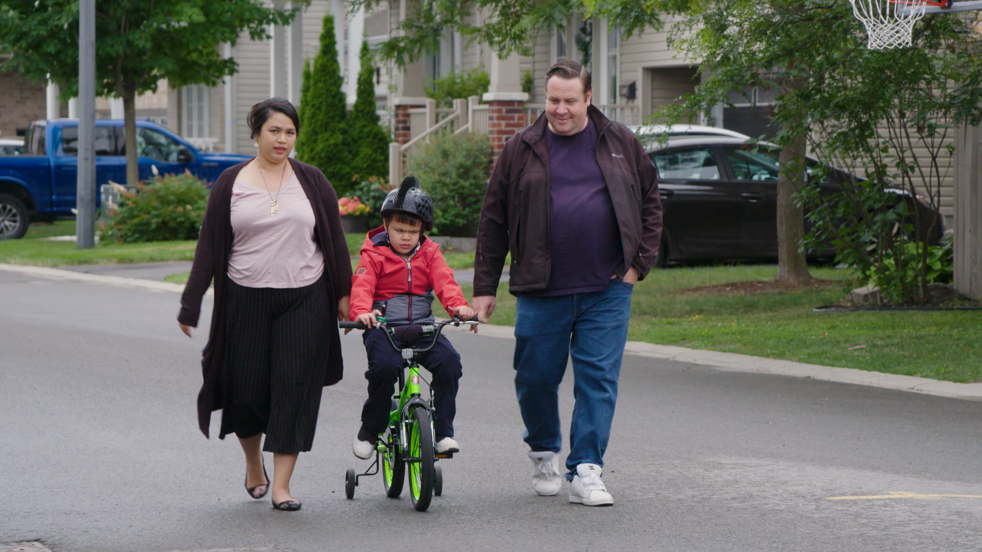 A mother and father stand on either side of their child are they bike on the street with training wheels and while wearing a helmet. 