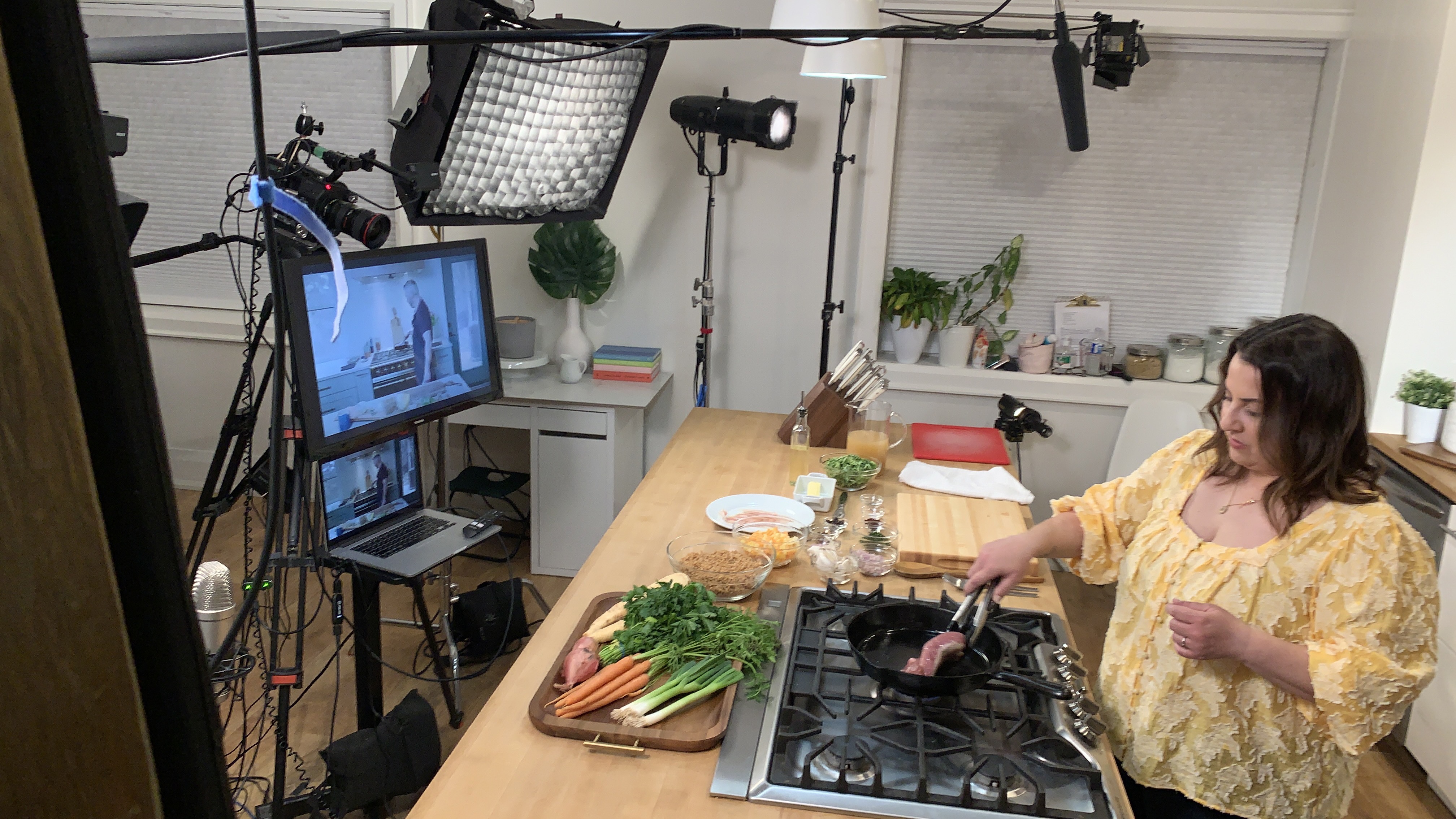 A photo of Mary Mammoliti cooking on the set of a kitchen with food on the table and cameras and lighting around.