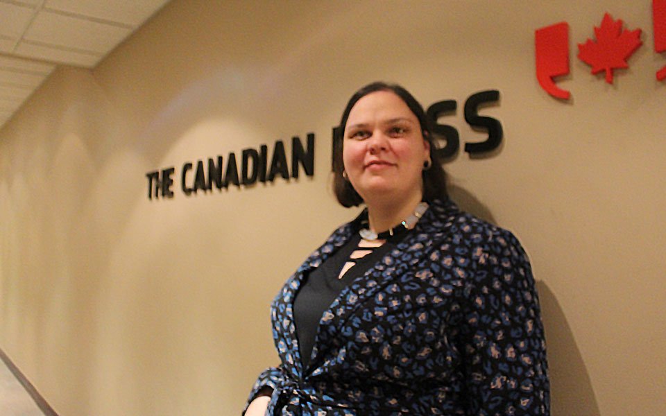 A photo of Michelle McQuigge smiling and leaning on a brown wall that has the Canadian Press logo. 