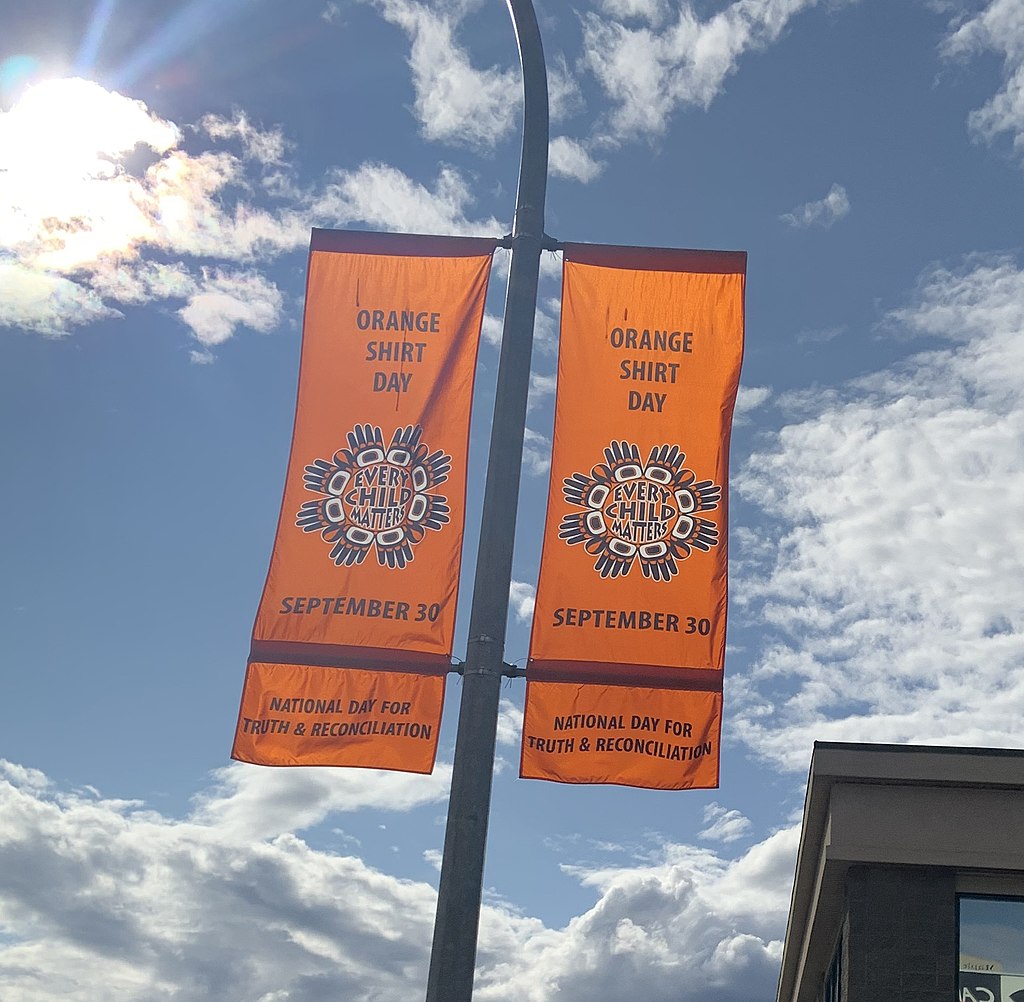 Two vertical Orange banners hang on a pole with the blue sly behind them. The banner reads "Every child matters, orange shirt day, September 30. National Day for Truth and Reconciliation."